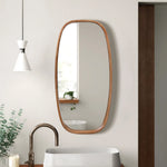 A modern wall mirror with an oval wooden frame hangs on a white wall above the sink, accompanied by a white pendant light and a planter brimming with greenery.