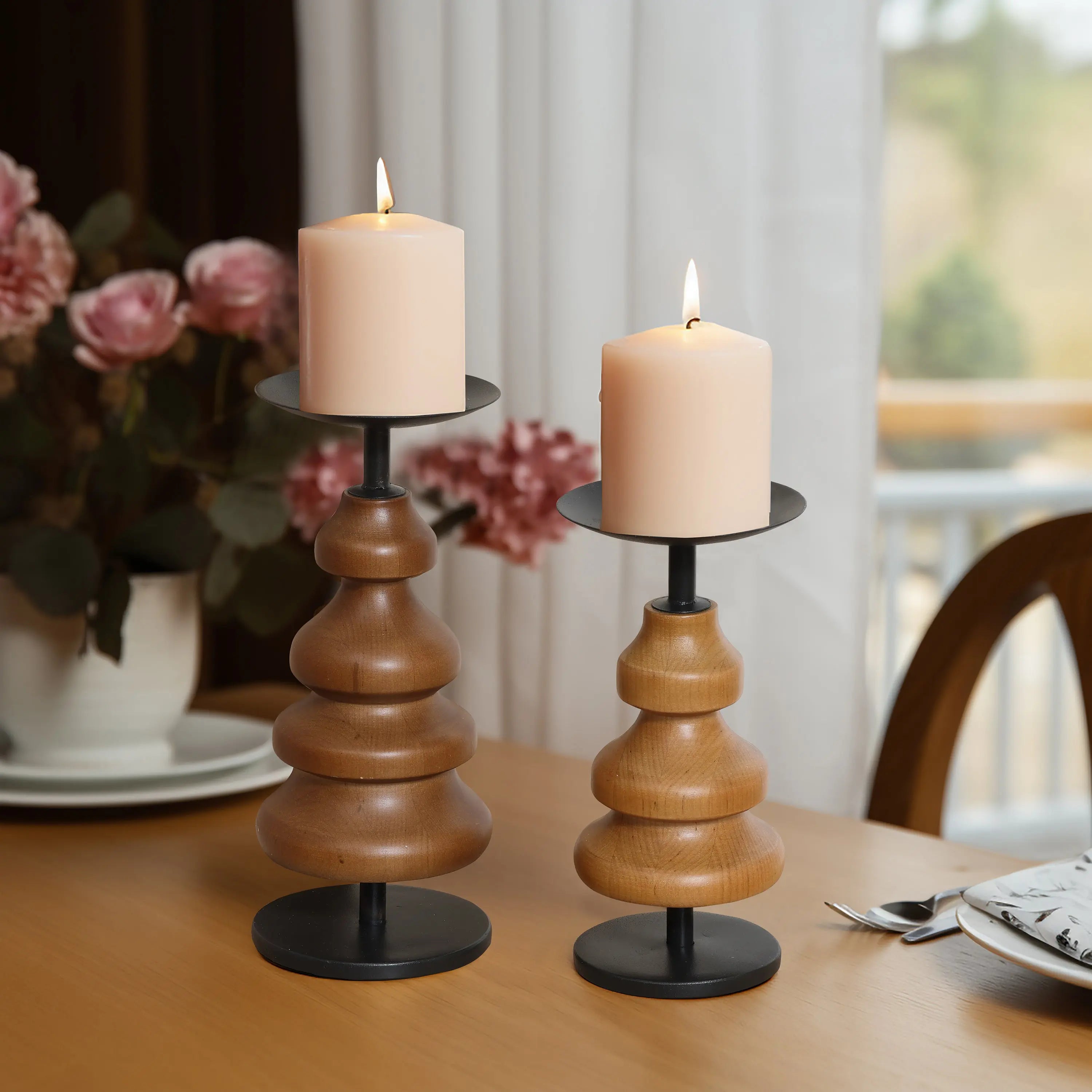 Two lit pillar candles rest on elegant birch wood holders, enhancing the dining table with a warm glow, while pink flowers bloom softly in the background, creating an exquisite home decor setting.