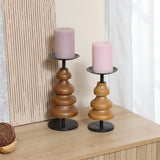 Two birch wood candle holders with black metal bases, each cradling a pink pillar candle, grace a wooden surface near a textured panel and a window with sheer curtains, adding serene elegance to your home decor.