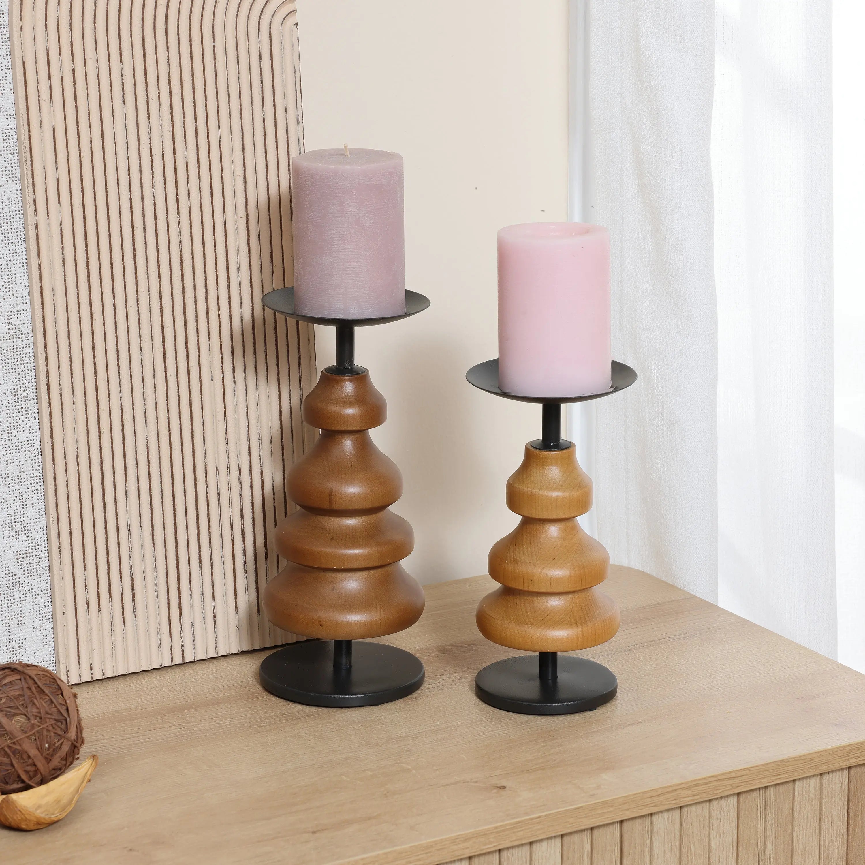 Two birch wood candle holders with black metal bases, each cradling a pink pillar candle, grace a wooden surface near a textured panel and a window with sheer curtains, adding serene elegance to your home decor.