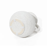 A speckled white ceramic mug with a wide body and a single handle, reminiscent of a decorative jug design, rests upside down on a white surface.