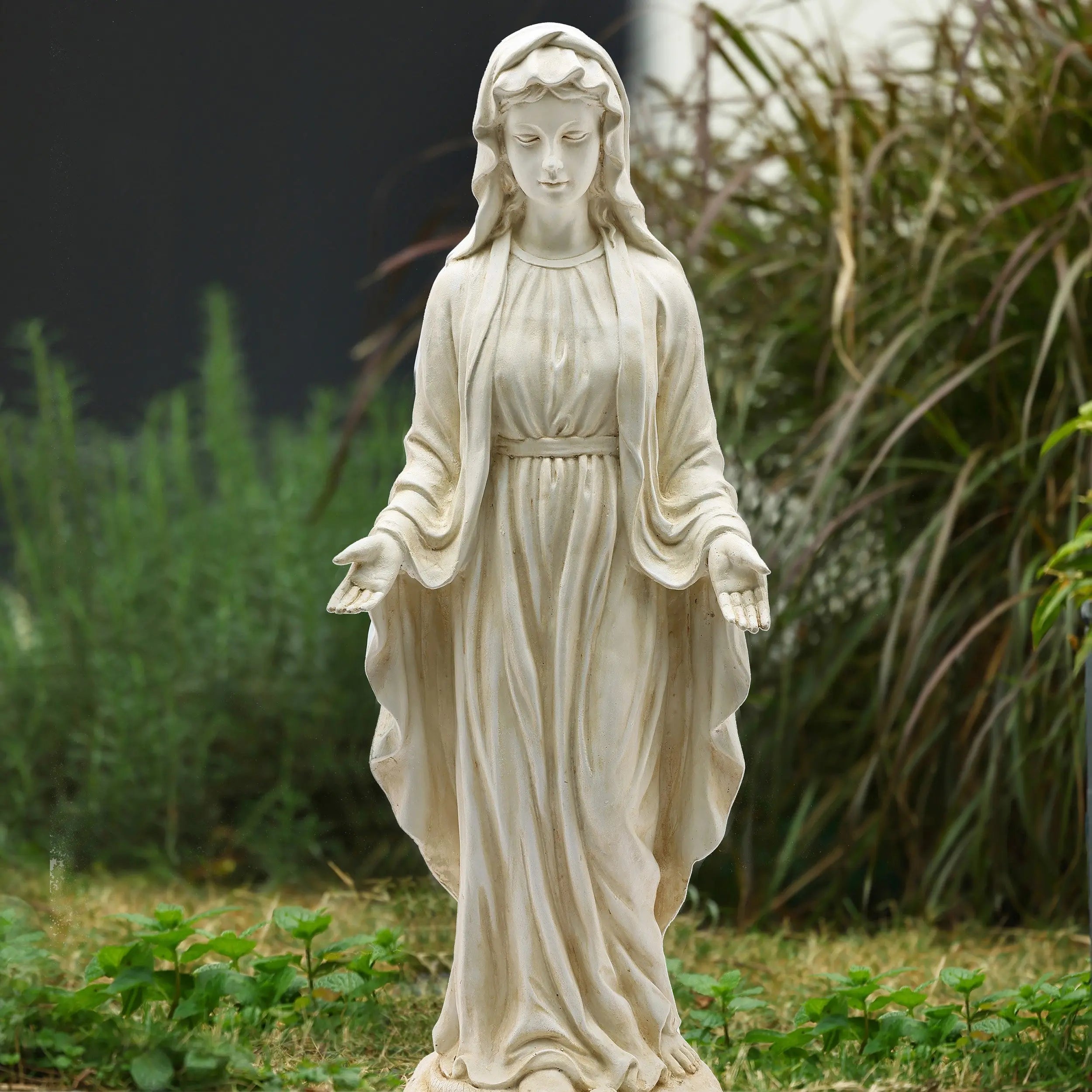 A white Mary statue with a vintage ivory finish, depicted in flowing robes and outstretched hands, stands outdoors surrounded by green plants and grass.
