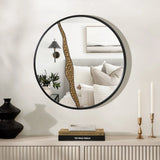 Round mirror with a black frame reflects a bedroom with pillows and a wall sconce. The left half of the mirror features an artistic design with a decorative golden crack. Two candles and books enhance the modern decoration on the console table.