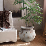 A rustic room features a LuxenHome Antique cauldron planter with a tall green plant on a woven rug. A beige armchair with a brown blanket is to the left, while a painting and soft lighting enhance the vintage rustic look, creating an inviting atmosphere.
