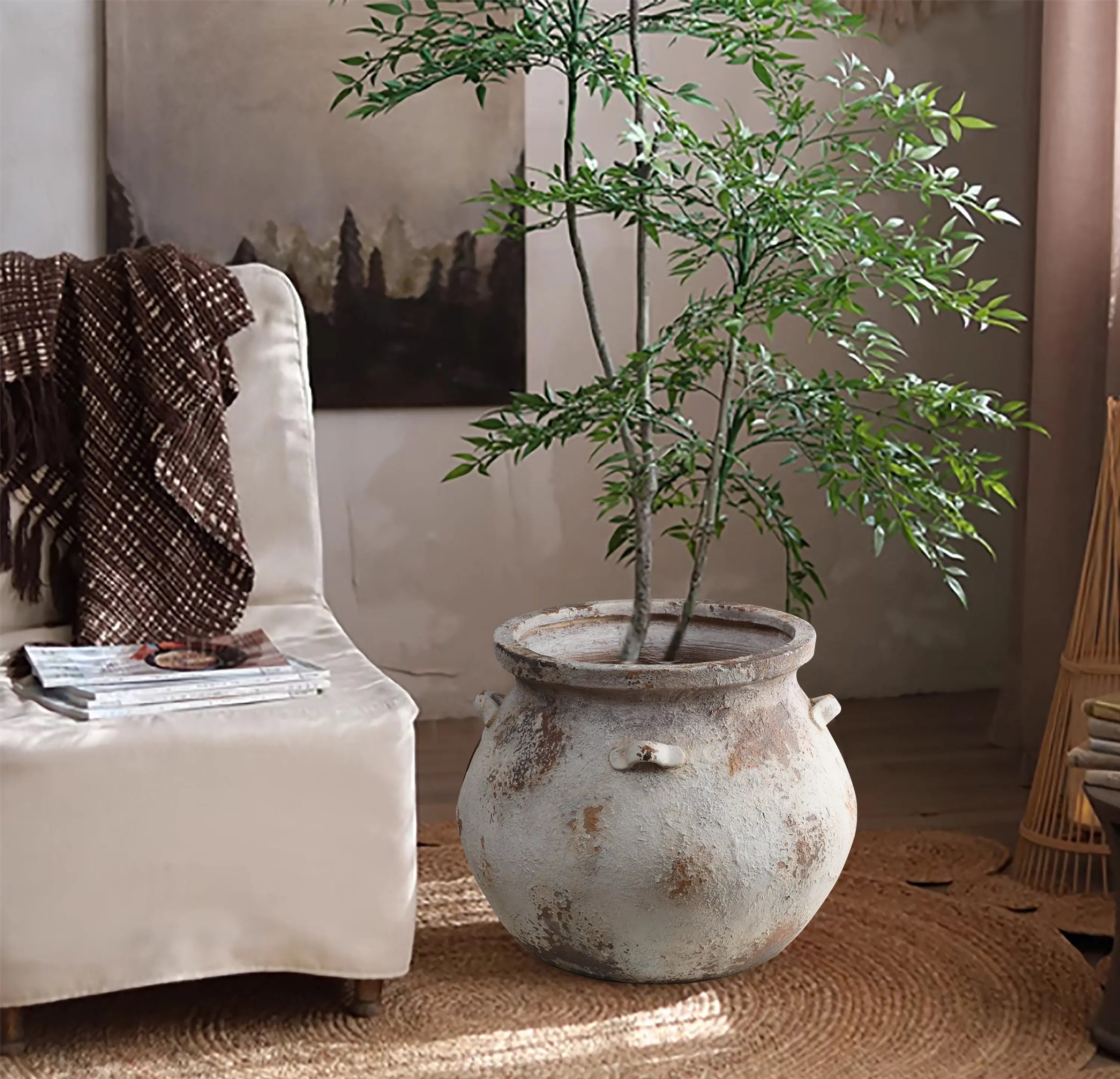 A rustic room features a LuxenHome Antique cauldron planter with a tall green plant on a woven rug. A beige armchair with a brown blanket is to the left, while a painting and soft lighting enhance the vintage rustic look, creating an inviting atmosphere.