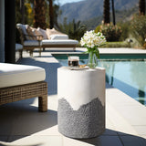 A round patio table with a marble top and textured base holds a glass vase of white flowers and a drink. Positioned next to a white cushioned chair near the swimming pool, this charming setup offers elegant outdoor seating for relaxation.