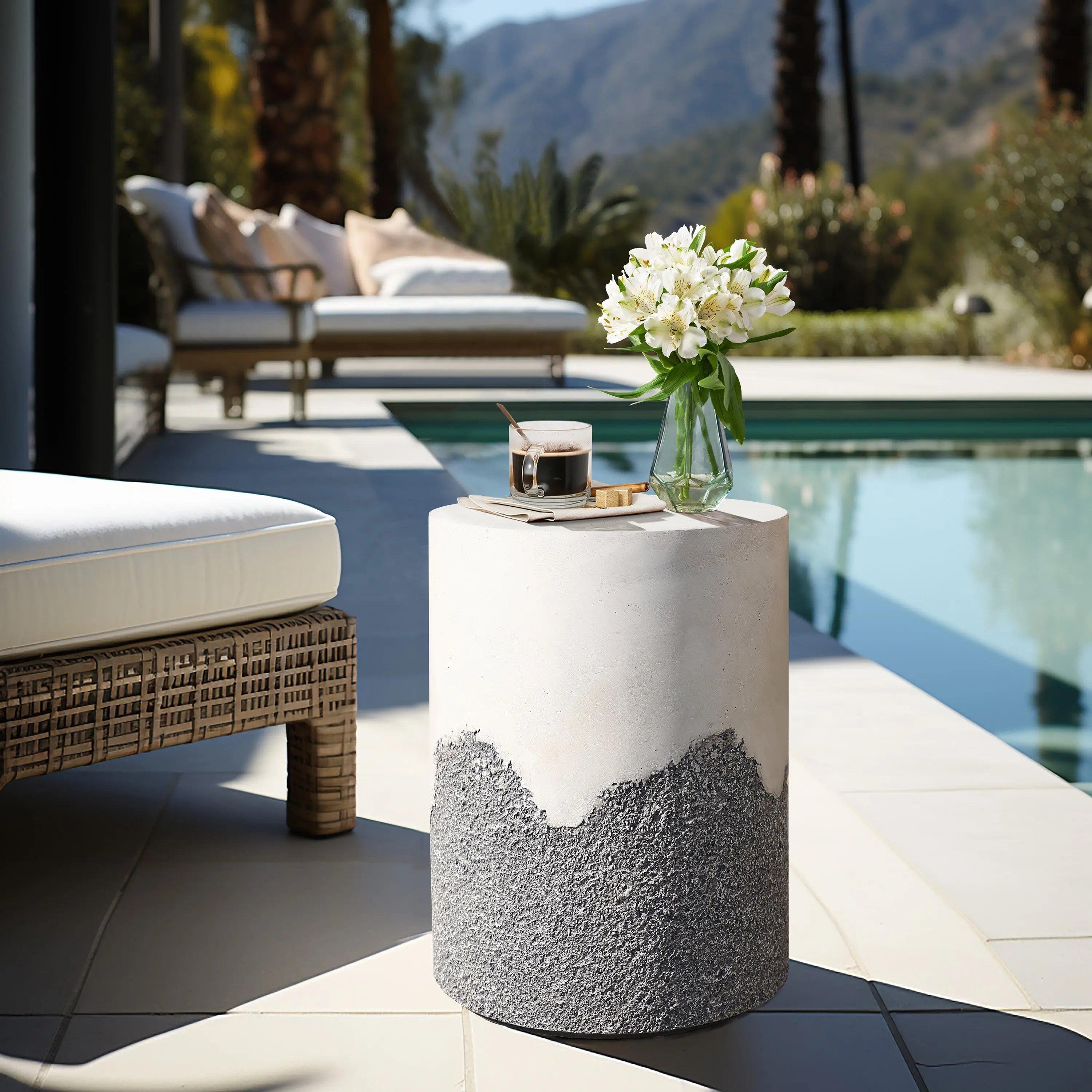 A round patio table with a marble top and textured base holds a glass vase of white flowers and a drink. Positioned next to a white cushioned chair near the swimming pool, this charming setup offers elegant outdoor seating for relaxation.