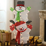 Two lighted snowmen, stacked one on top of the other, brighten the scene as part of a charming Lighted Snowman Set with gift boxes nearby and a cozy fireplace visible on the right—perfect for your outdoor holiday decorations.