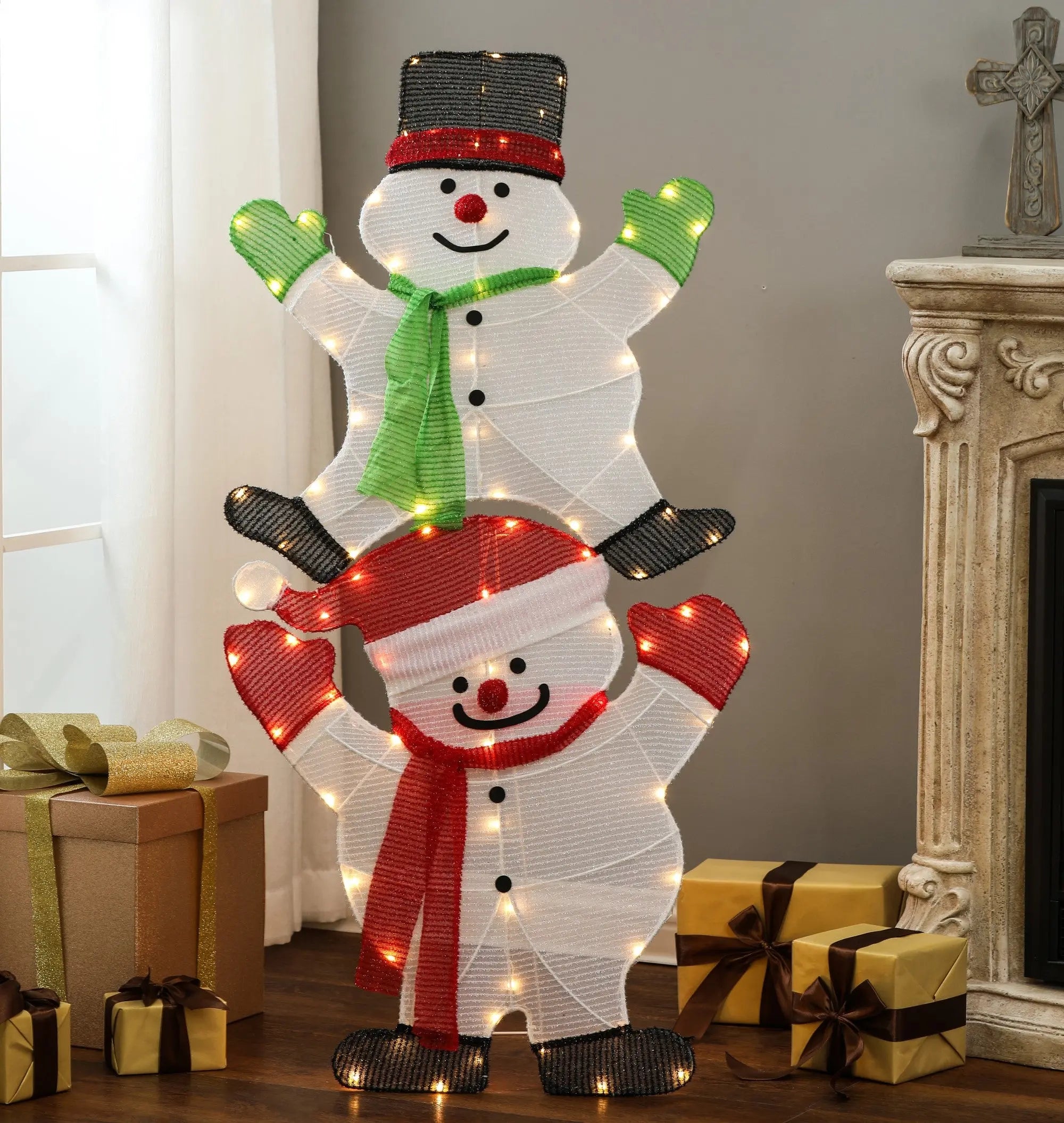 Two lighted snowmen, stacked one on top of the other, brighten the scene as part of a charming Lighted Snowman Set with gift boxes nearby and a cozy fireplace visible on the right—perfect for your outdoor holiday decorations.