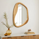 A uniquely shaped wooden mirror is mounted on a white wall above a wooden console, creating an organic vibe. A vase with greenery and two small ceramic decorations add natural decor to the scene.