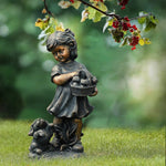 A whimsical statue, this bronze girl holding a basket stands on grass beside her small dog, with tree branches and red berries overhead—the perfect weather-resistant addition to any garden statues collection.