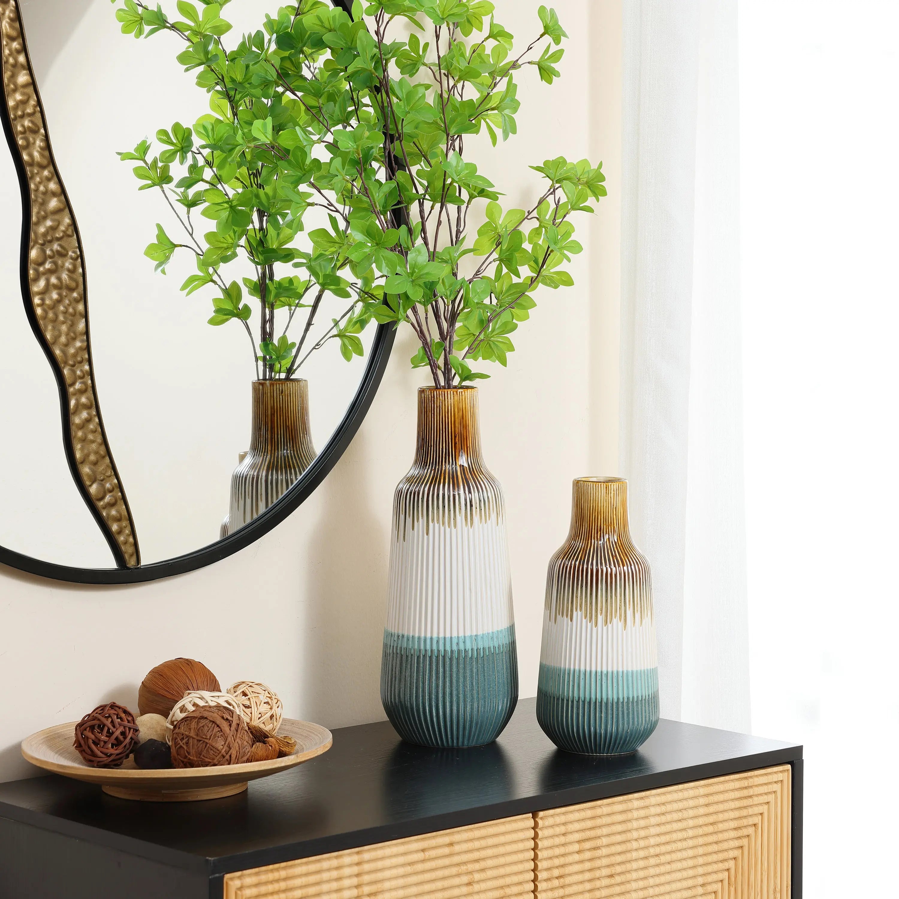 Two high-quality ceramic vases with green branches sit on a wooden cabinet next to a round mirror. A decorative bowl with assorted spheres adds charm to this stylish home decor arrangement.