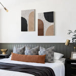 The bedroom boasts a neatly made bed with a brown leather bolster, patterned pillows, and a dark throw. Above it, abstract wall art featuring geometric shapes adds visual intrigue and sophistication to the space.