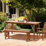 A solid Malaysian hardwood outdoor dining table set with a bench and chair graces the stone patio, surrounded by lush trees and plants. The table, treated with teak oil coating, features a vase of flowers and a tray, creating an inviting natural ambiance.