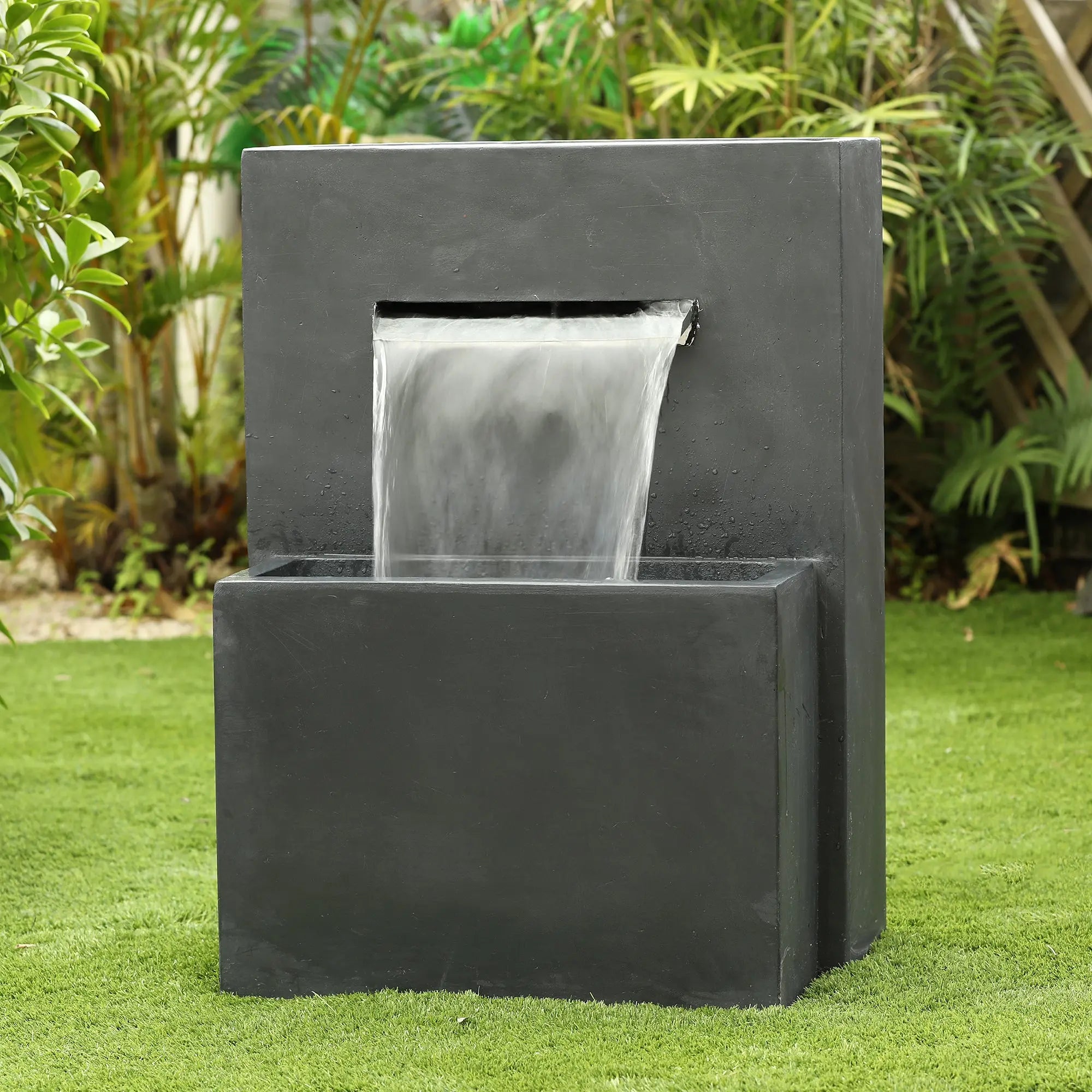 A modern gray rectangular waterfall outdoor fountain with built-in LED lighting features water flowing from an upper spout into a lower basin, set on green grass with lush plants in the background.
