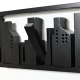 This abstract black metal sculpture of a city skyline, featuring rectangular buildings, serves as a unique wall-mounted coat rack. Crafted from solid fir wood with flip-down hooks, it combines functionality and artistic flair on your white wall.