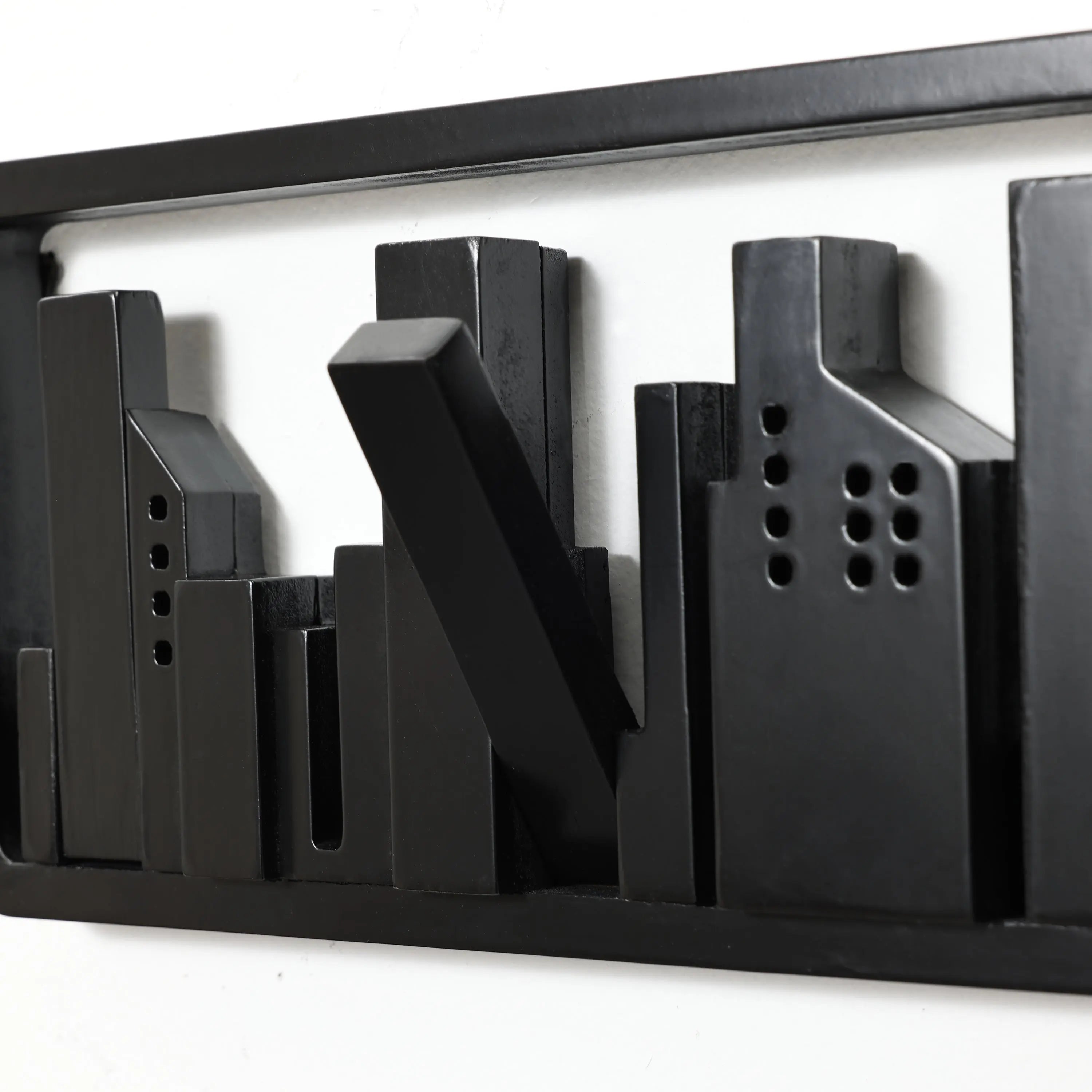 This abstract black metal sculpture of a city skyline, featuring rectangular buildings, serves as a unique wall-mounted coat rack. Crafted from solid fir wood with flip-down hooks, it combines functionality and artistic flair on your white wall.