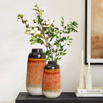 Two vintage ceramic vases with a color block gradient of orange and brown sit elegantly on a black table. One vase holds branches adorned with white flowers, lending charm to the scene. Nearby, two unlit candles complement the setup, while framed artwork graces the wall as perfect home decor.