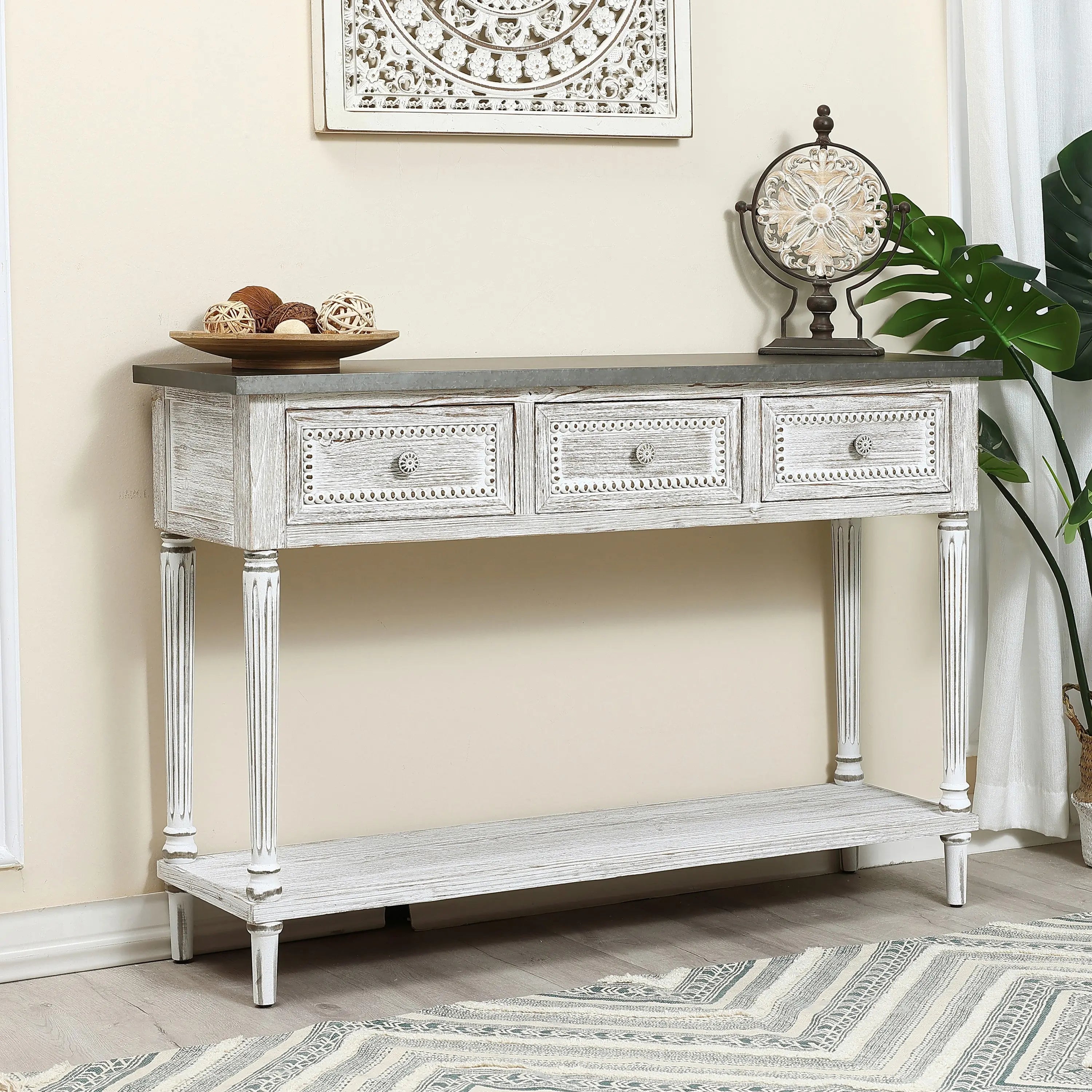 A farmhouse-inspired table, this white distressed wood console features three drawers, a lower shelf, and is adorned with potpourri, an ornate clock, and a large green plant nearby.