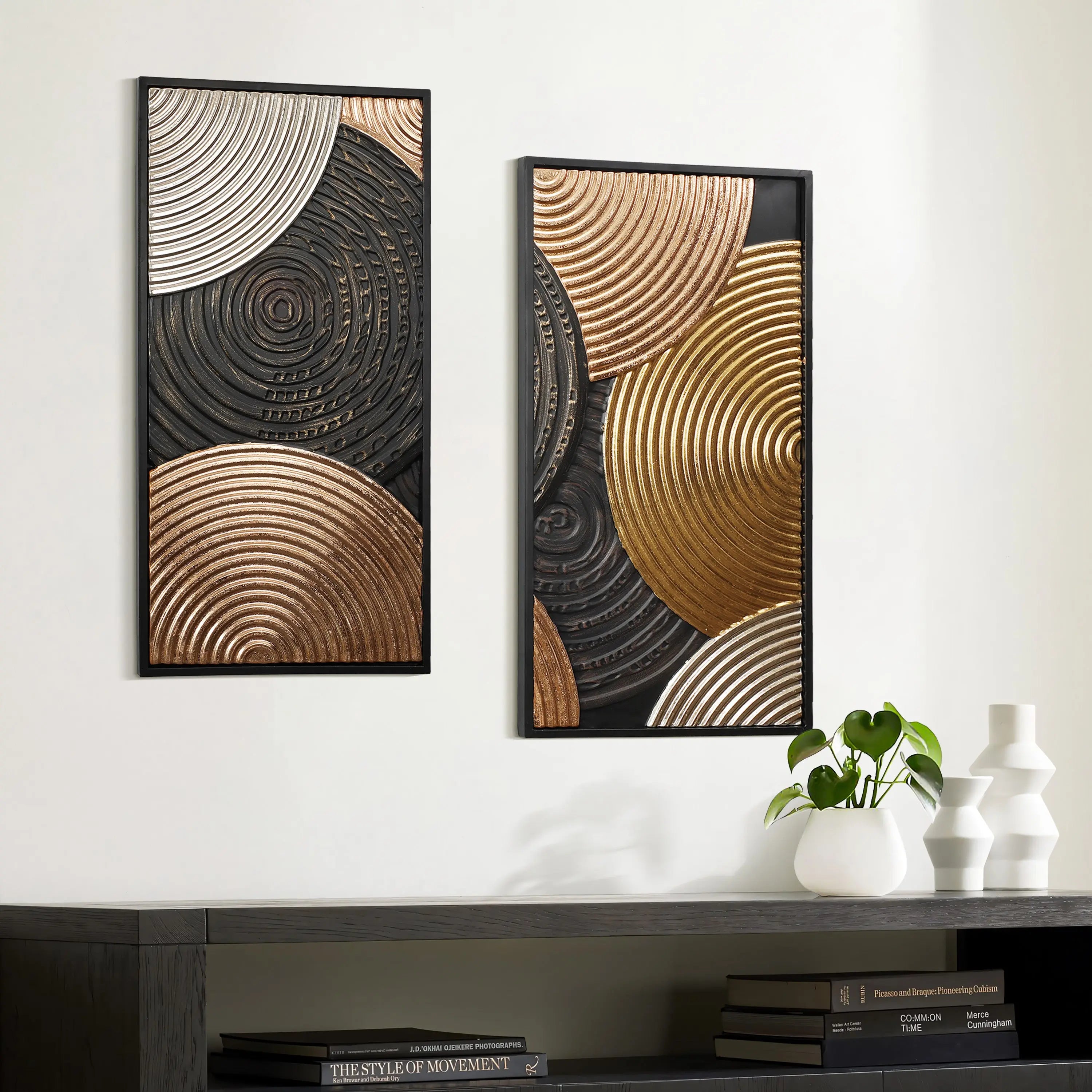 Two abstract art pieces with circular patterns in gold, black, and bronze tones adorn the white wall. Below is a dark table featuring a plant, books, and two white vases. The embossed textures of the frames enhance the overall elegance of this wall decor.