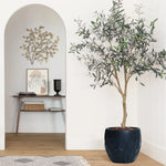 A geometric pot planter made of magnesium oxide material holds a potted plant, resting on a wooden floor beside a decorative wall. Artwork and a small table enhance the backdrop, perfect for indoor and outdoor use.