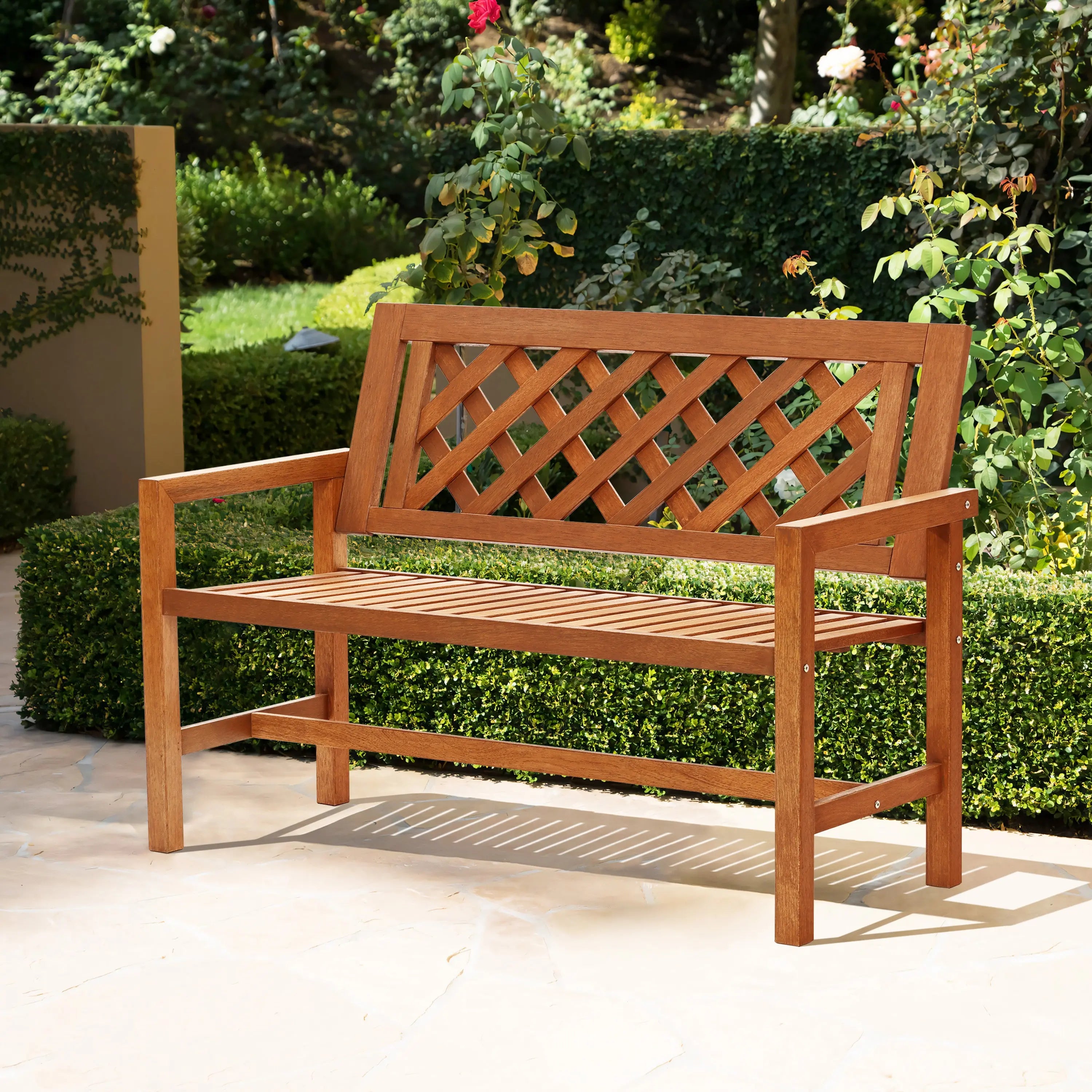 A solid wood garden bench with a lattice backrest adorns the stone patio, surrounded by lush green bushes and blooming roses.