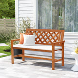 A garden bench with a lattice backrest features white and orange cushions, perfect for relaxing on the patios gray pavers. Crafted with solid wood construction, this outdoor bench sits against a backdrop of a white building and lush greenery.