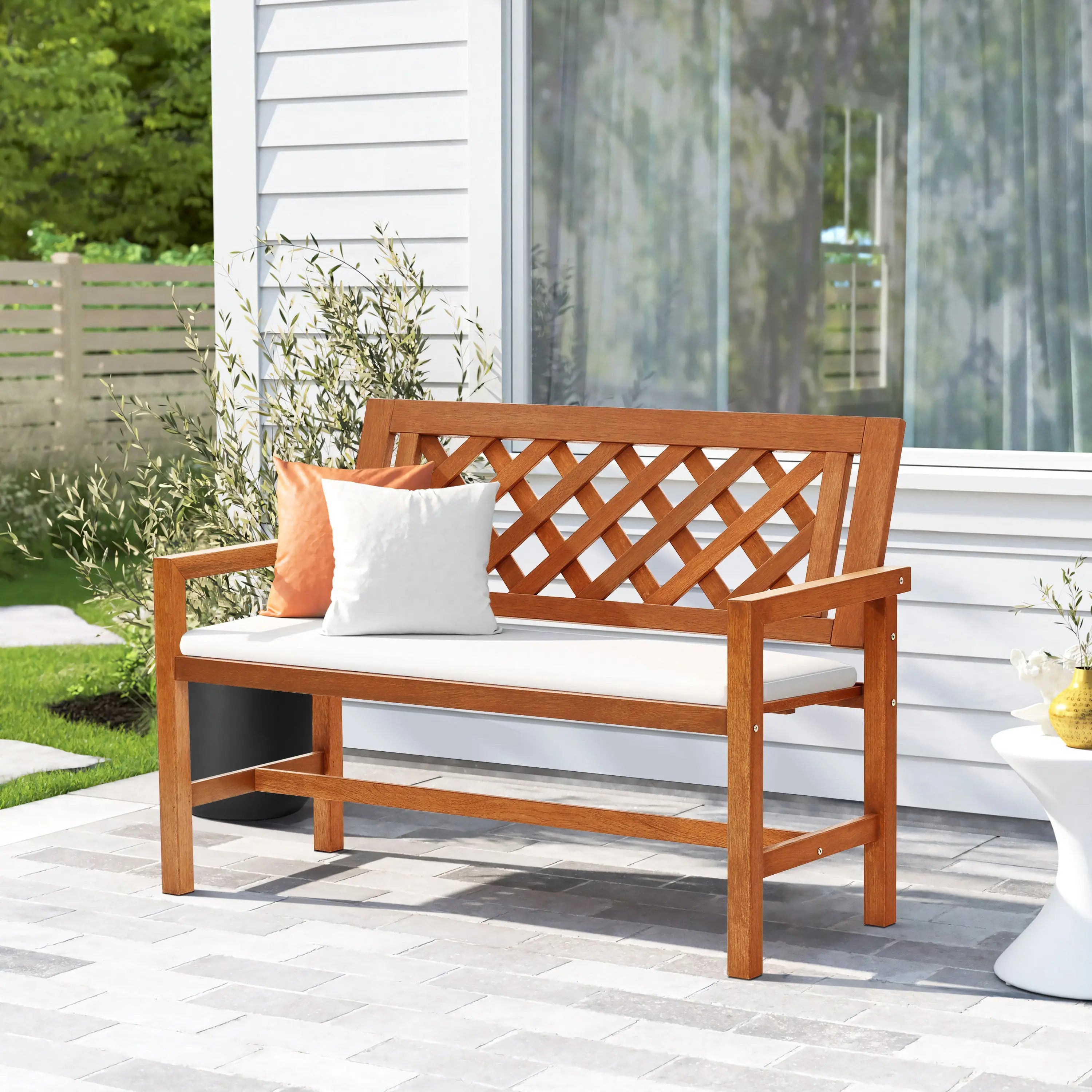 A garden bench with a lattice backrest features white and orange cushions, perfect for relaxing on the patios gray pavers. Crafted with solid wood construction, this outdoor bench sits against a backdrop of a white building and lush greenery.