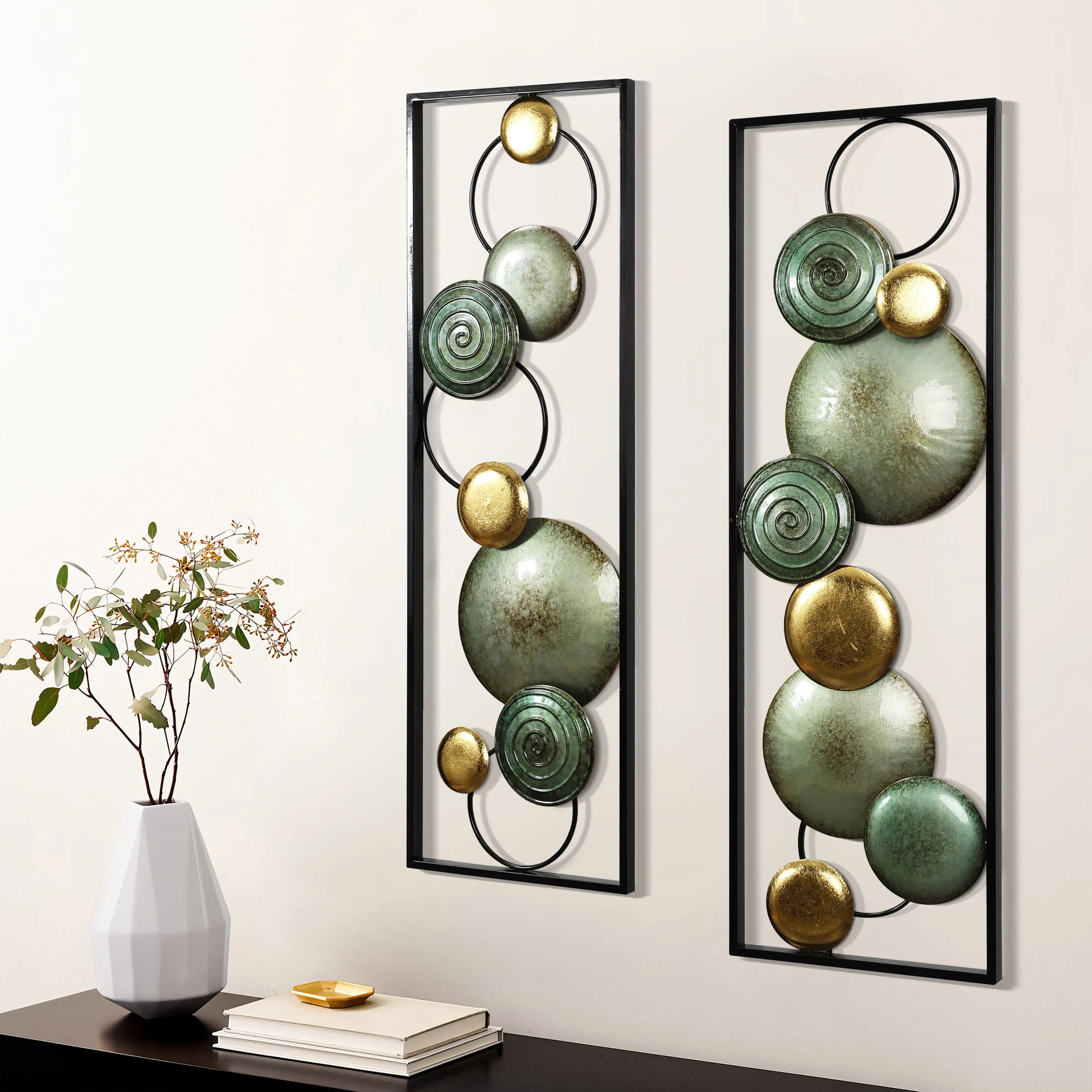 The LuxenHome Glam dishes metal wall art, set of 2, features circular teal and gold designs with metallic finishes. Perfect for a modern setting, they are displayed on a light wall beside a minimalist white vase with a delicate plant, enhancing the sleek decor.