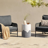 Two gray wicker chairs with cushions flank a cement side table with an ombre style finish, holding a vase of flowers and a cup. A beige throw and pillow add texture to the outdoor seating arrangement.