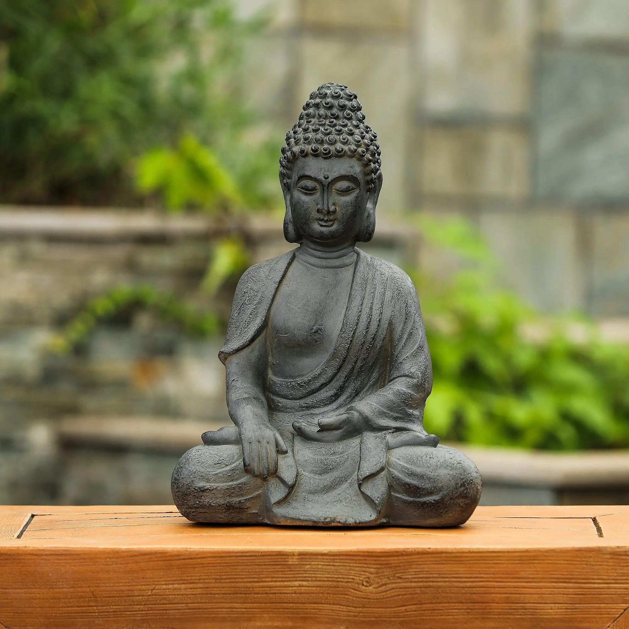 A weather resistant stone Buddha statue in a meditative pose rests on a wooden surface, surrounded by greenery and a stone wall, perfect for serene garden decor.