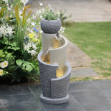 A modern tiered stone flower pot water fountain with a small potted plant on top, surrounded by flowers and greenery, set on a tiled outdoor area—perfect for adding elegance to garden spaces.