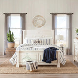 A bright bedroom features a white bed with striped bedding, a jute rug, potted plants, and two windows with sheer curtains. Above the bed, abstract wall decor in a gold tone finish adds an elegant touch.