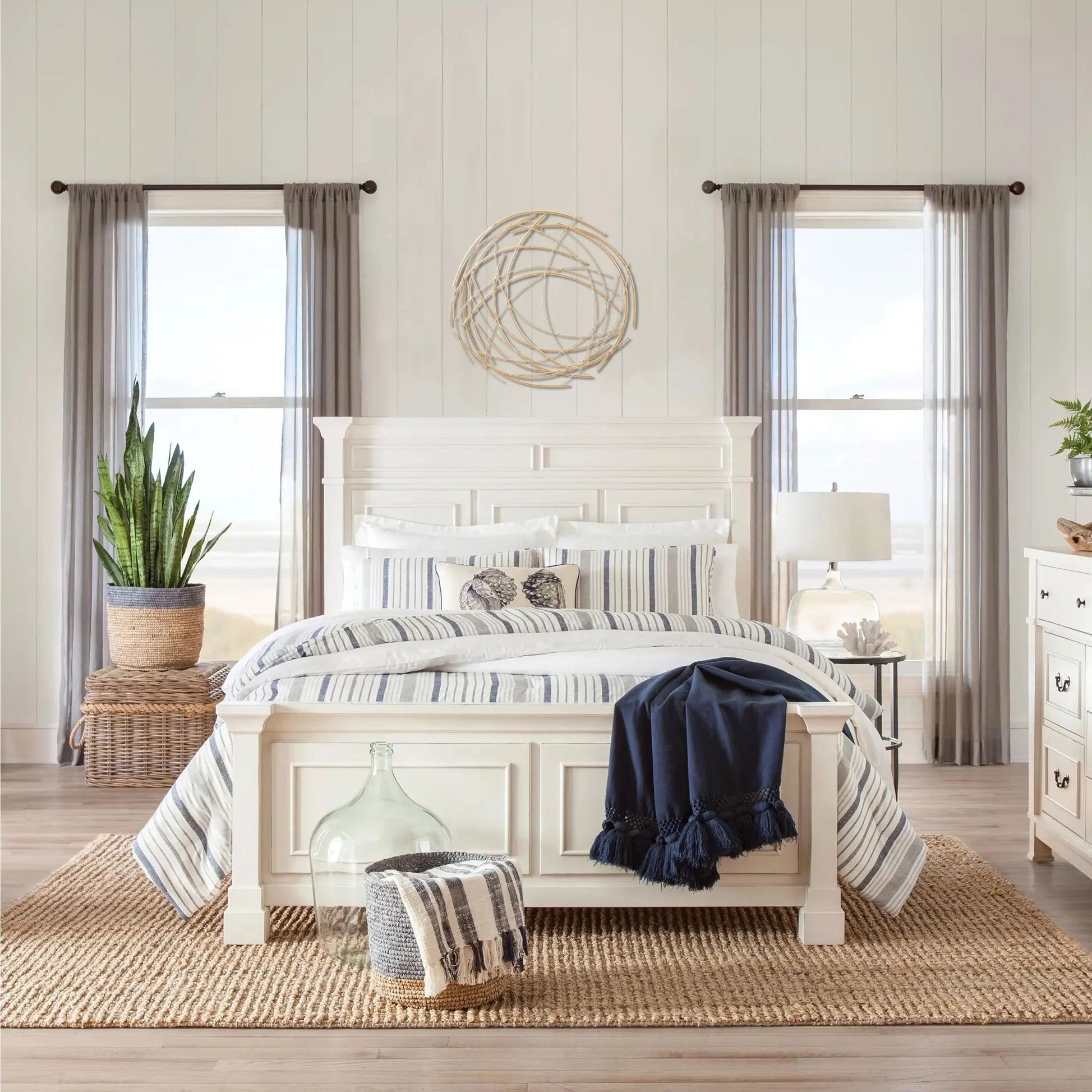 A bright bedroom features a white bed with striped bedding, a jute rug, potted plants, and two windows with sheer curtains. Above the bed, abstract wall decor in a gold tone finish adds an elegant touch.
