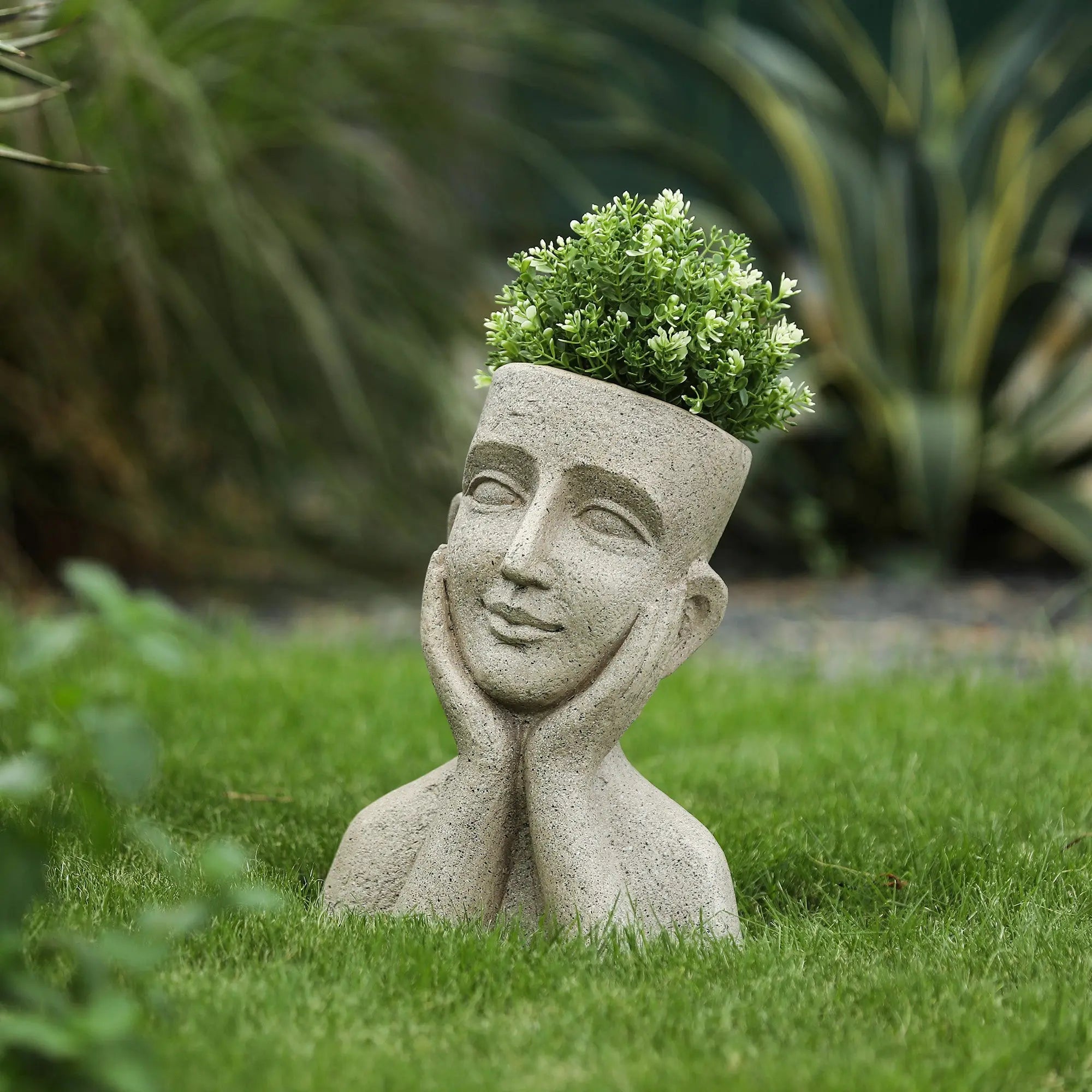 A bust head planter shaped like a human head and hands rests on grass, with green foliage growing from the top like hair.