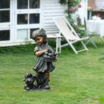 Whimsical statue of a bronze-colored girl holding a basket with a small dog at her feet, placed on a grassy lawn near a white building and outdoor furniture—perfect for adding charm to your garden décor.