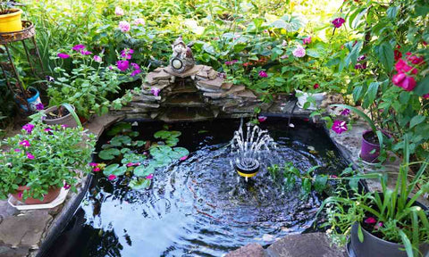 An outdoor fountain in a small pond. It is surrounded by lush green plants, purple flowers, and a small yard statue.