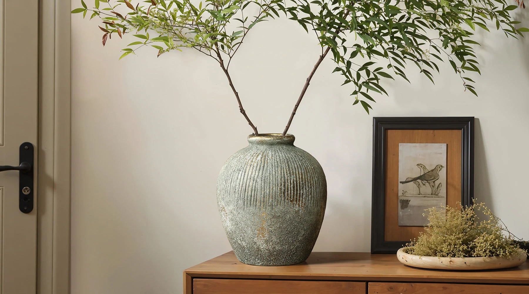 A large, textured gray-green vase with eucalyptus branches sits on a wooden floor next to a wooden cabinet and a stack of books.