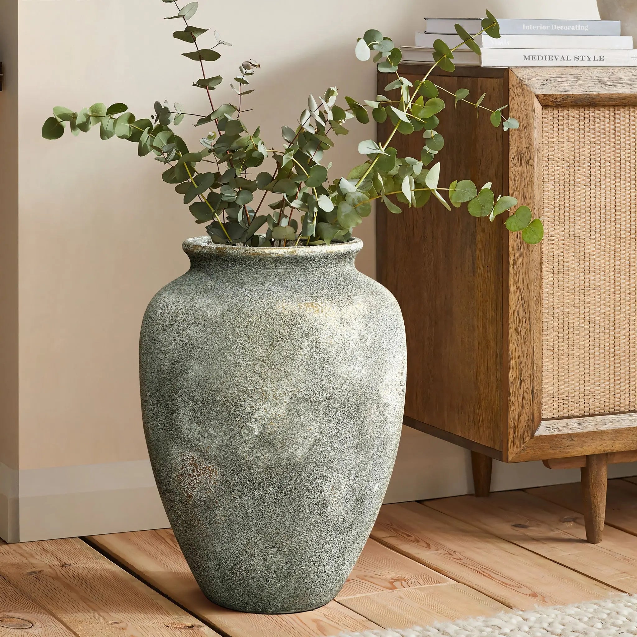 A large, textured gray-green vase with eucalyptus branches sits on a wooden floor next to a wooden cabinet and a stack of books.