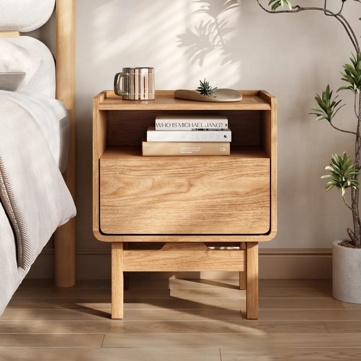 Wooden bedside table with two shelves, holding a striped mug, a book titled Who is Michael Jiang?, a small plant, and a smooth stone. A potted plant stands nearby on a wooden floor.