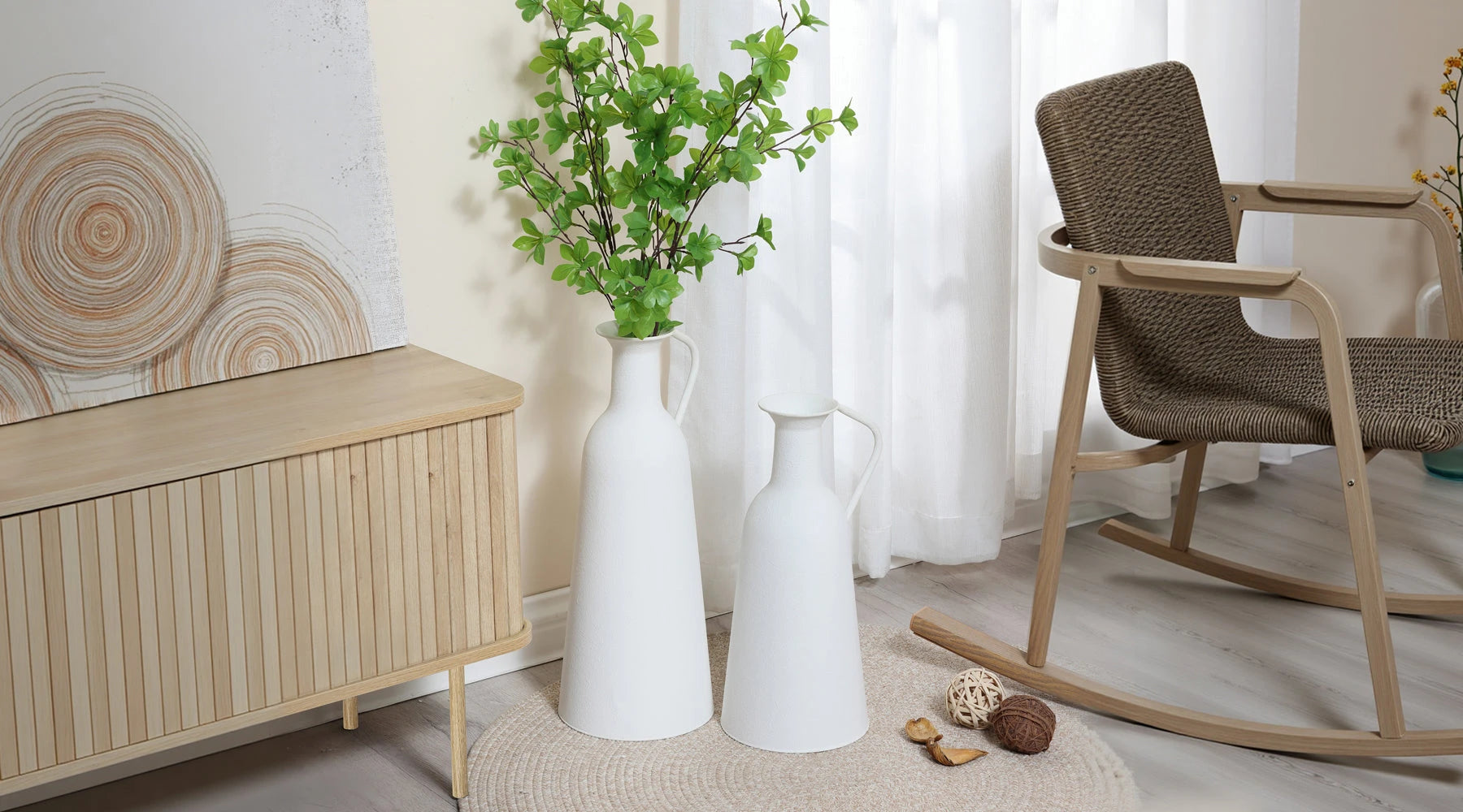 Two white ceramic vases, one tall with green foliage and one shorter, placed on the floor next to a wooden cabinet in a sunlit room with neutral decor.