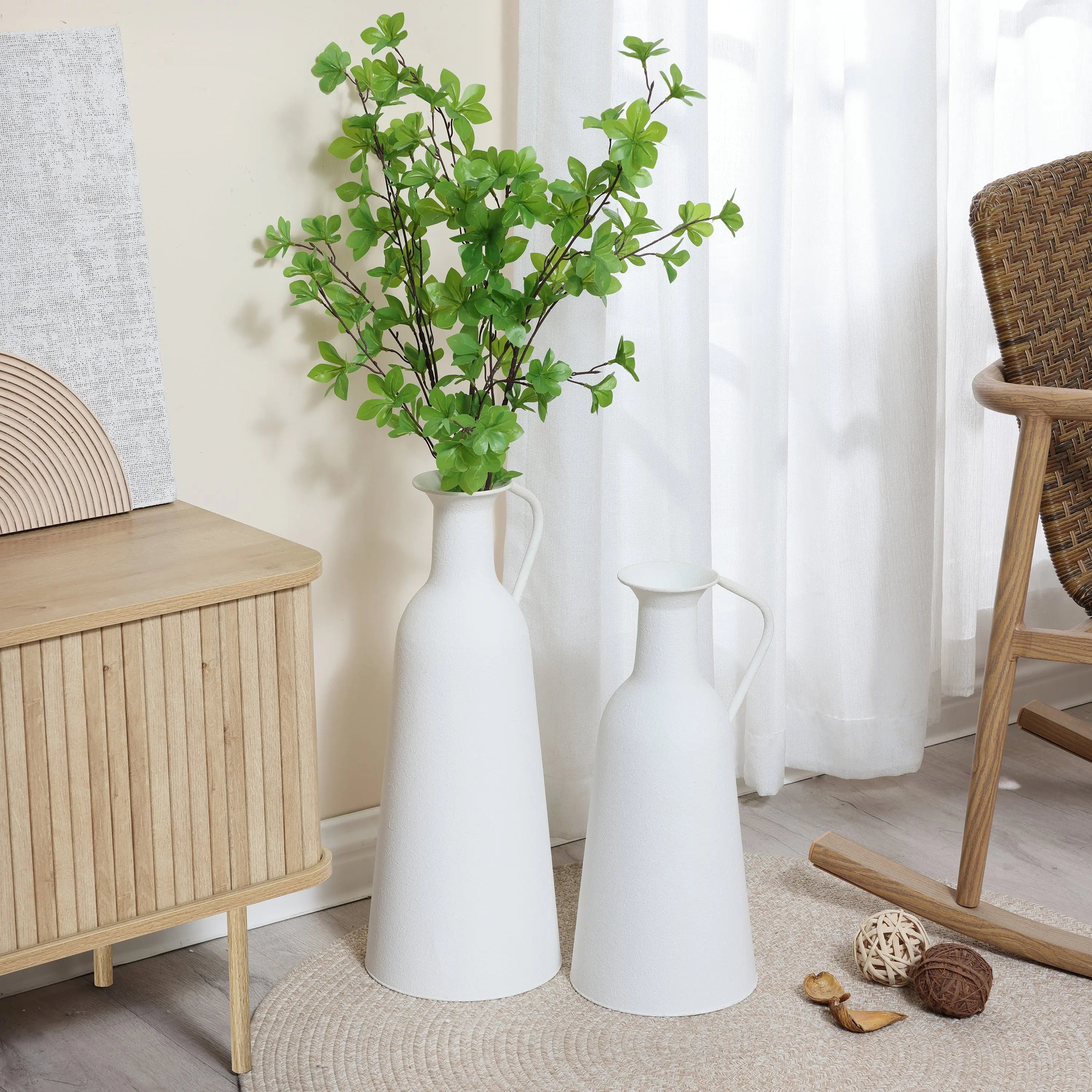 Two white ceramic vases, one tall with green foliage and one shorter, placed on the floor next to a wooden cabinet in a sunlit room with neutral decor.