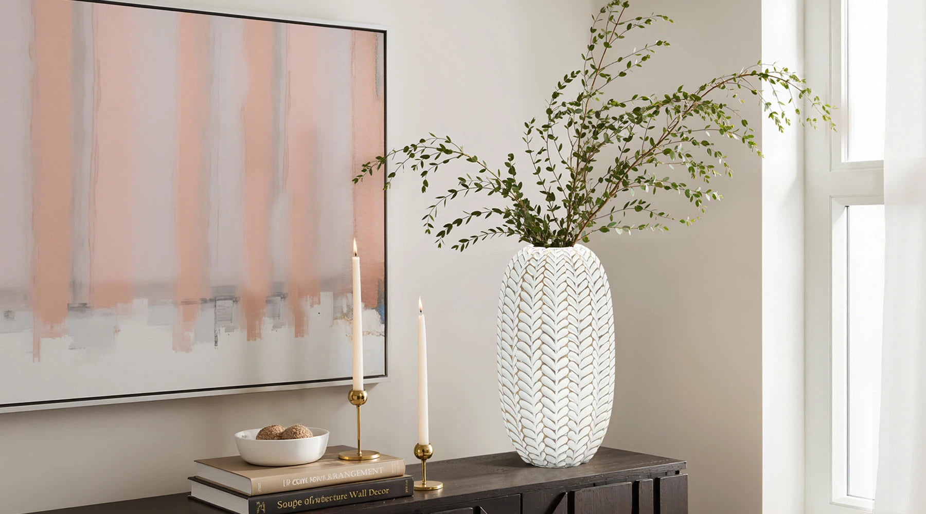 A tall, patterned vase with white artificial flowers is placed on a wooden table, next to round decorative objects in a well-lit, elegant room.