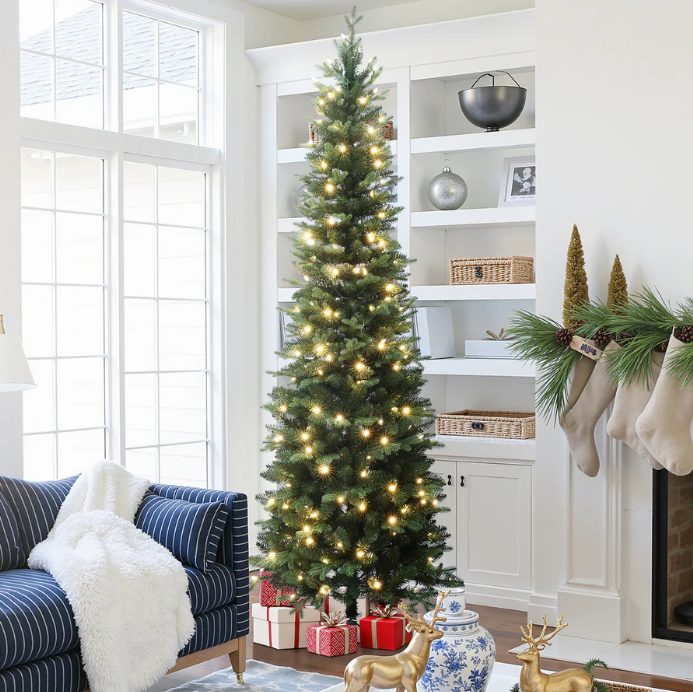 A decorated Christmas tree with lights and presents stands near a staircase adorned with garland and bows in a bright, modern living space.