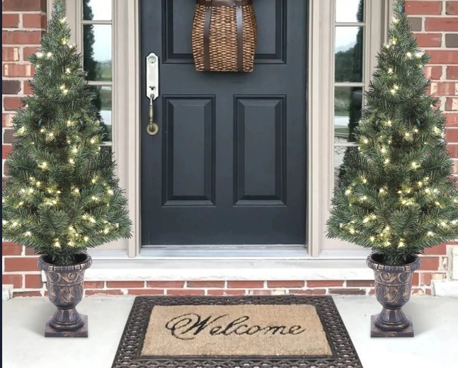 A snow-flocked Christmas tree with lights and red presents stands by a window in a cozy living room with a cream armchair and winter landscape painting on the wall.