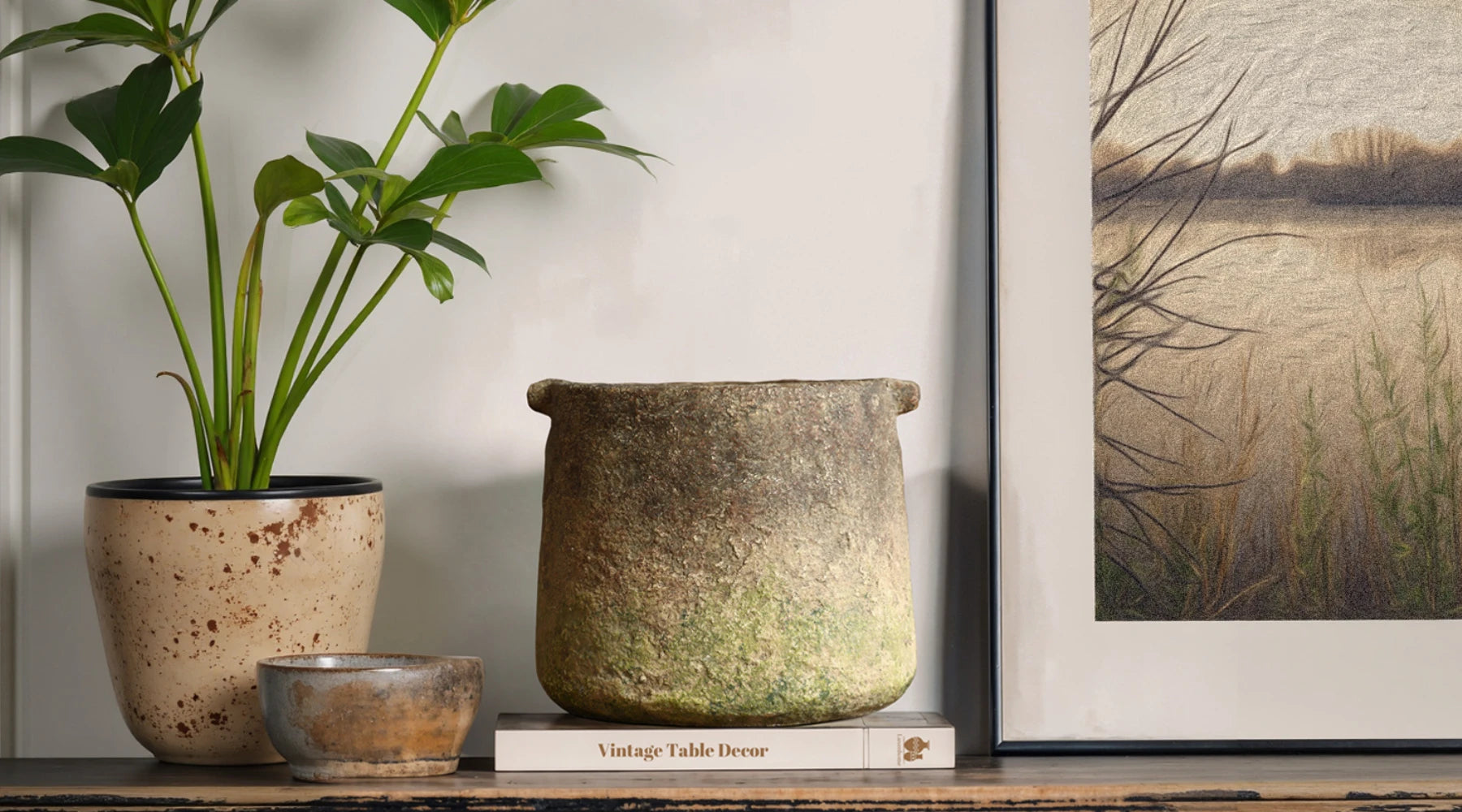 A rustic stone pot sits on a book titled Vintage Table Decor on a distressed wooden table, next to a small plant and framed artwork.