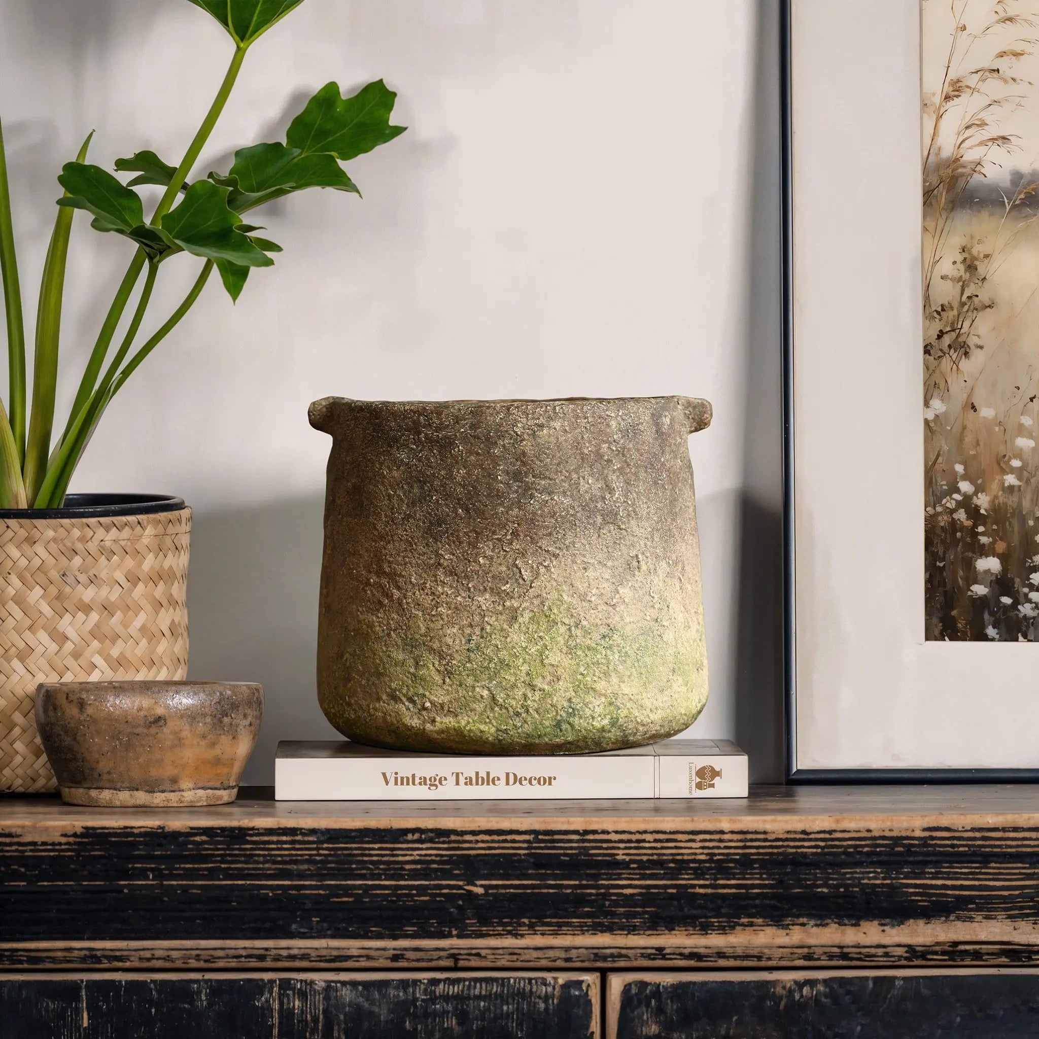 A rustic stone pot sits on a book titled Vintage Table Decor on a distressed wooden table, next to a small plant and framed artwork.