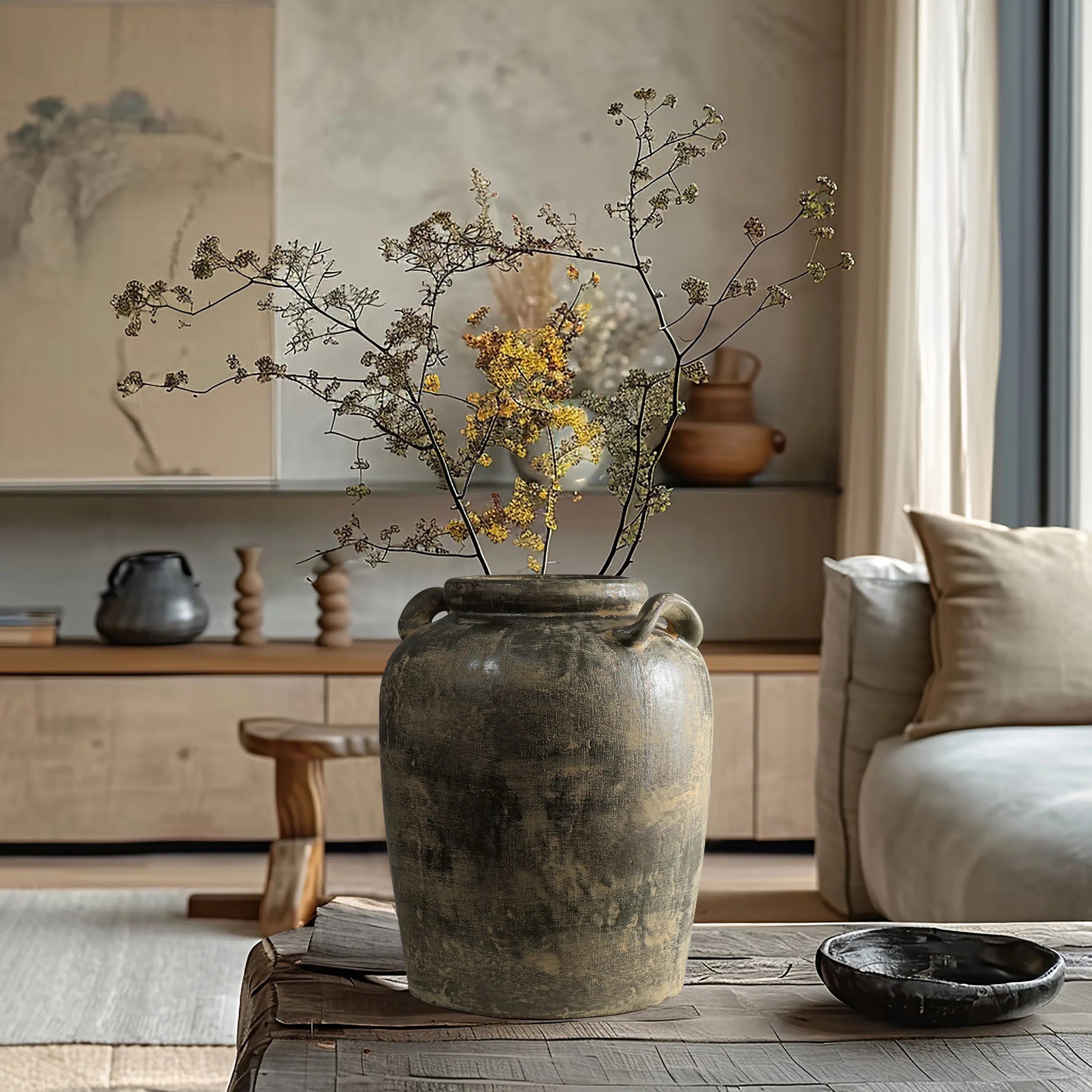 A large rustic ceramic vase with dried branches sits on a wooden table in a neutral-toned living room with minimalist decor.