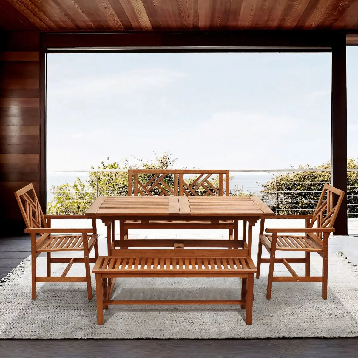 Outdoor wooden dining set with four chairs and a bench on a gray rug. The scene overlooks a deck with a view of a blue sky and distant water.