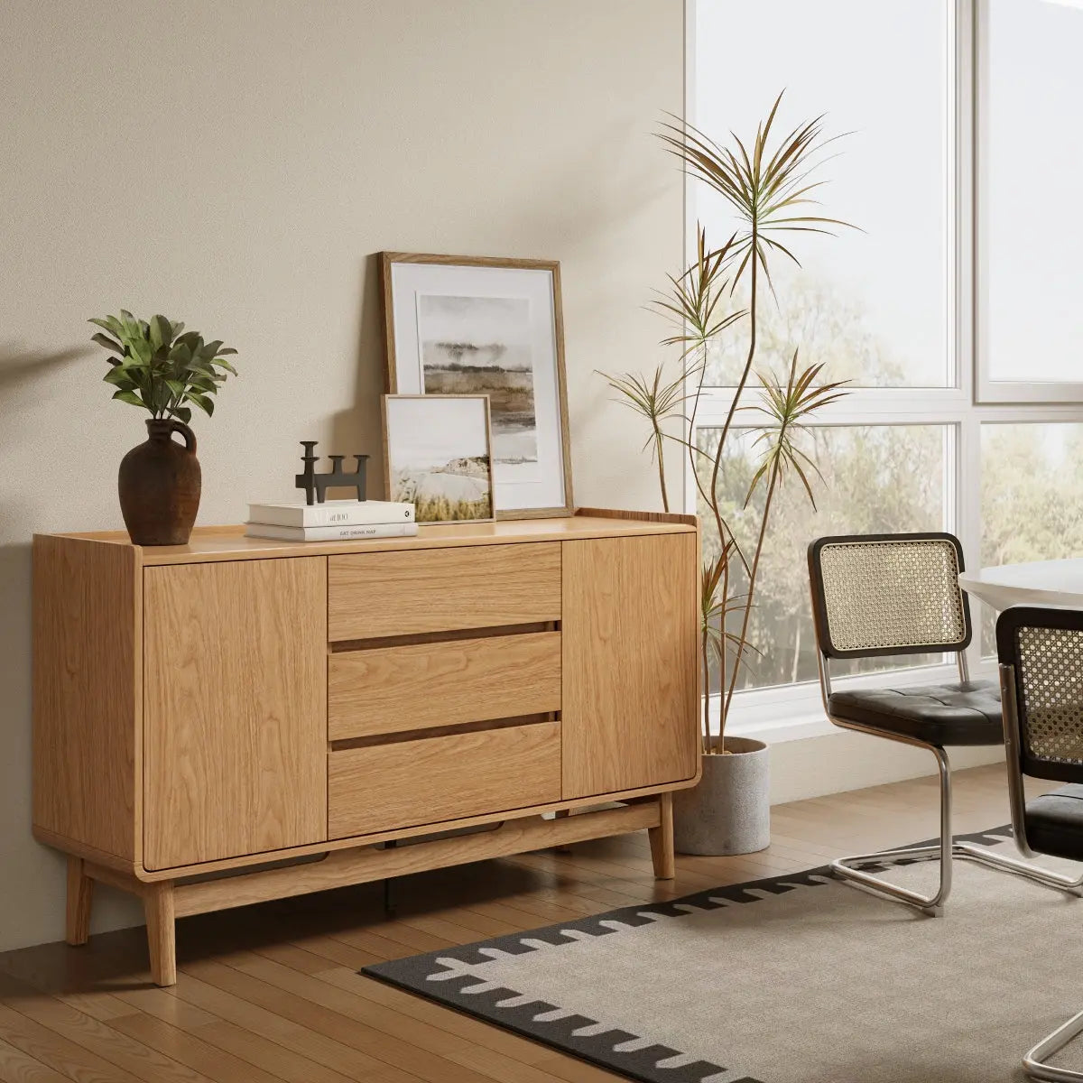 A wooden sideboard with plants, framed art, and decor stands beside two modern chairs and a large window in a bright, minimalist room.